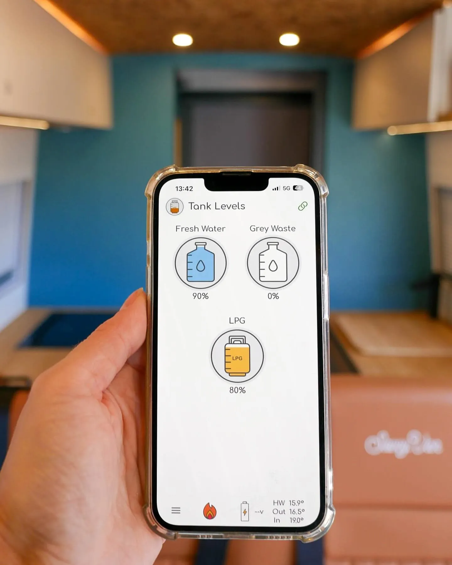 A hand holding a phone inside a campervan, showing the SavvyVan app’s tank levels: fresh water 90%, grey waste 0% and LPG 80%