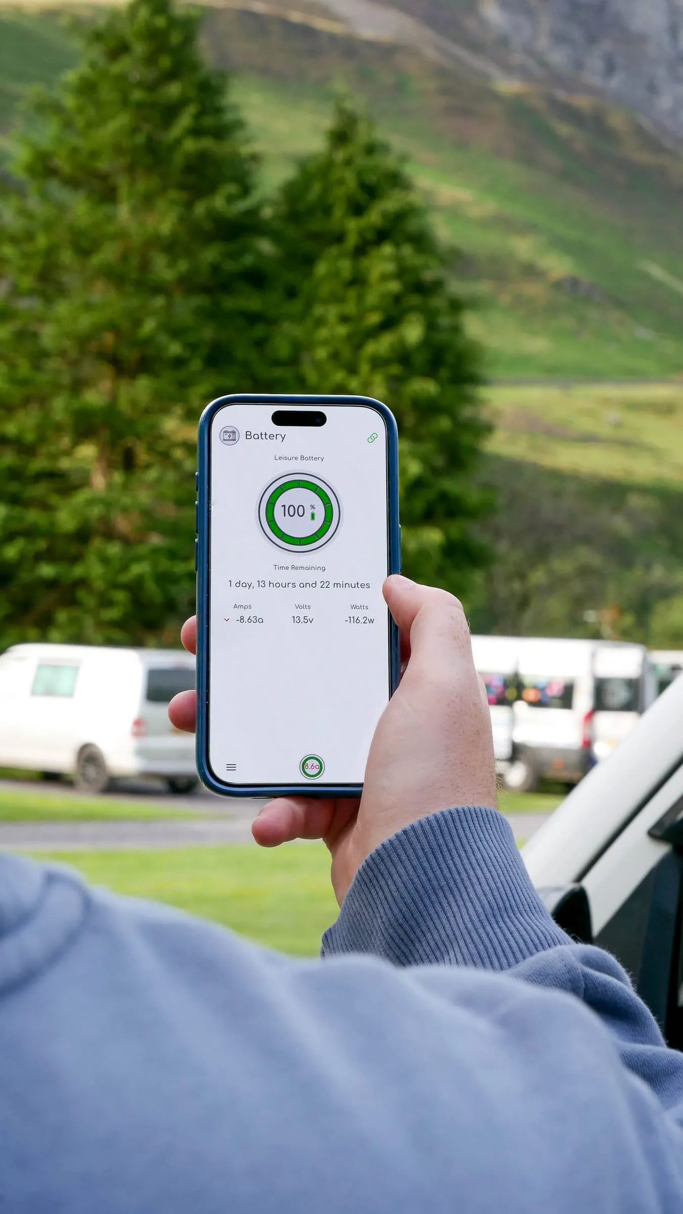 A person holding a phone showing the SavvyVan battery screen at 100 percent, with campervans parked on grass and a green hillside behind