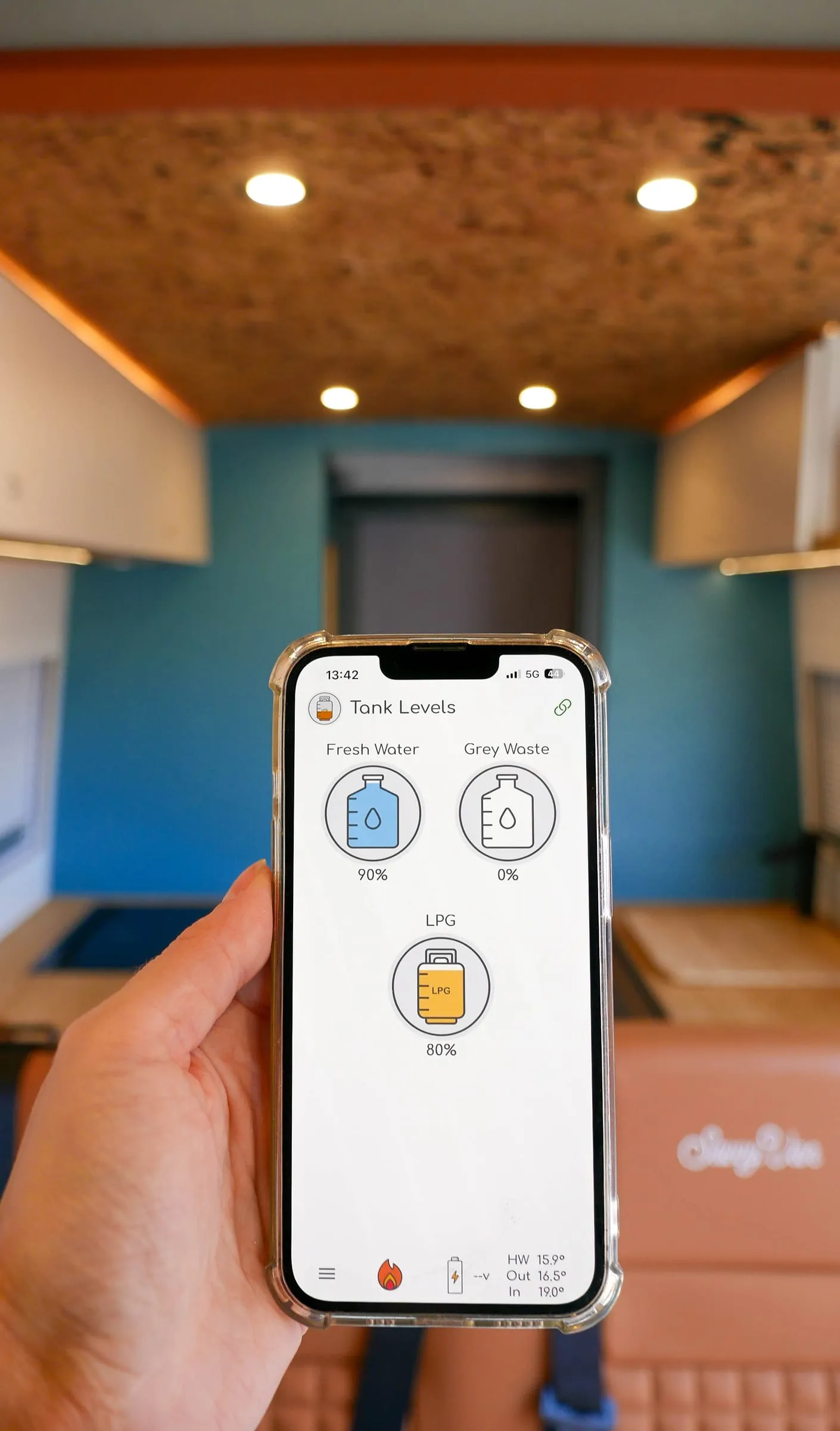 A phone showing the SavvyVan tank levels screen - fresh water 90 percent, grey waste 0 percent, LPG 80 percent - held up inside a finished campervan conversion