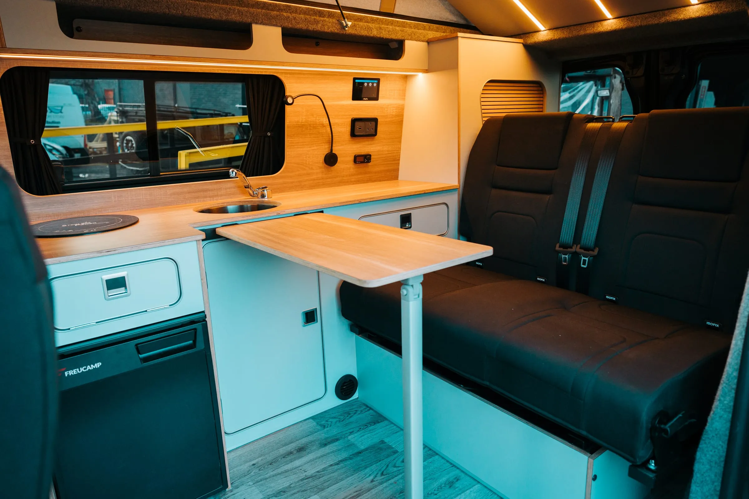 A finished campervan conversion with a SavvyVan3 panel on the oak splashback above the kitchen worktop, beside a reading light and a mains socket, with a rear bench seat and a fold-out table