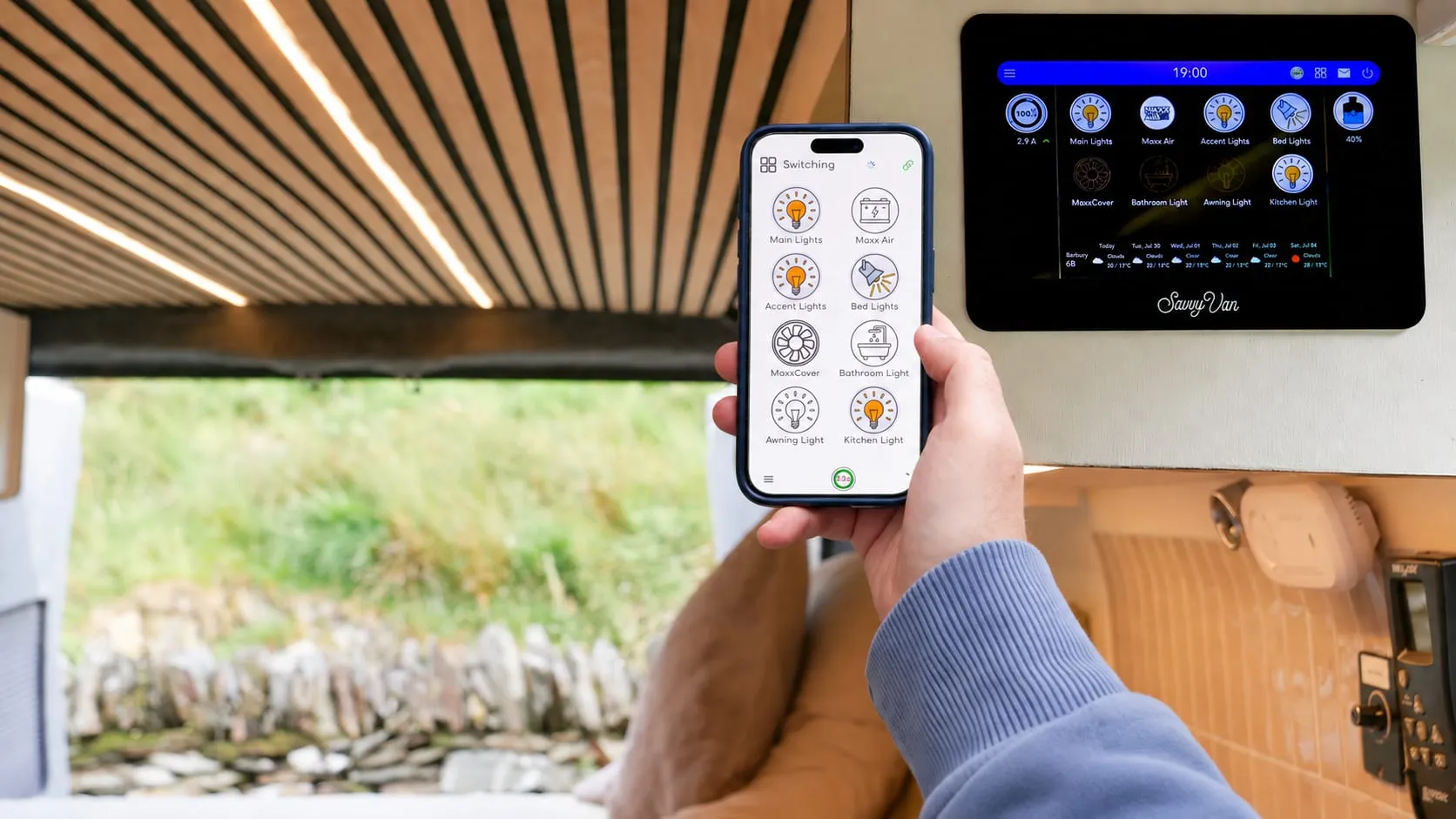 Inside a campervan with the side door open onto a stone wall and grass: a hand holding a phone showing the SavvyVan app's switching screen, with the same lights and fan on the SavvyVan3 panel fitted to the wall behind it