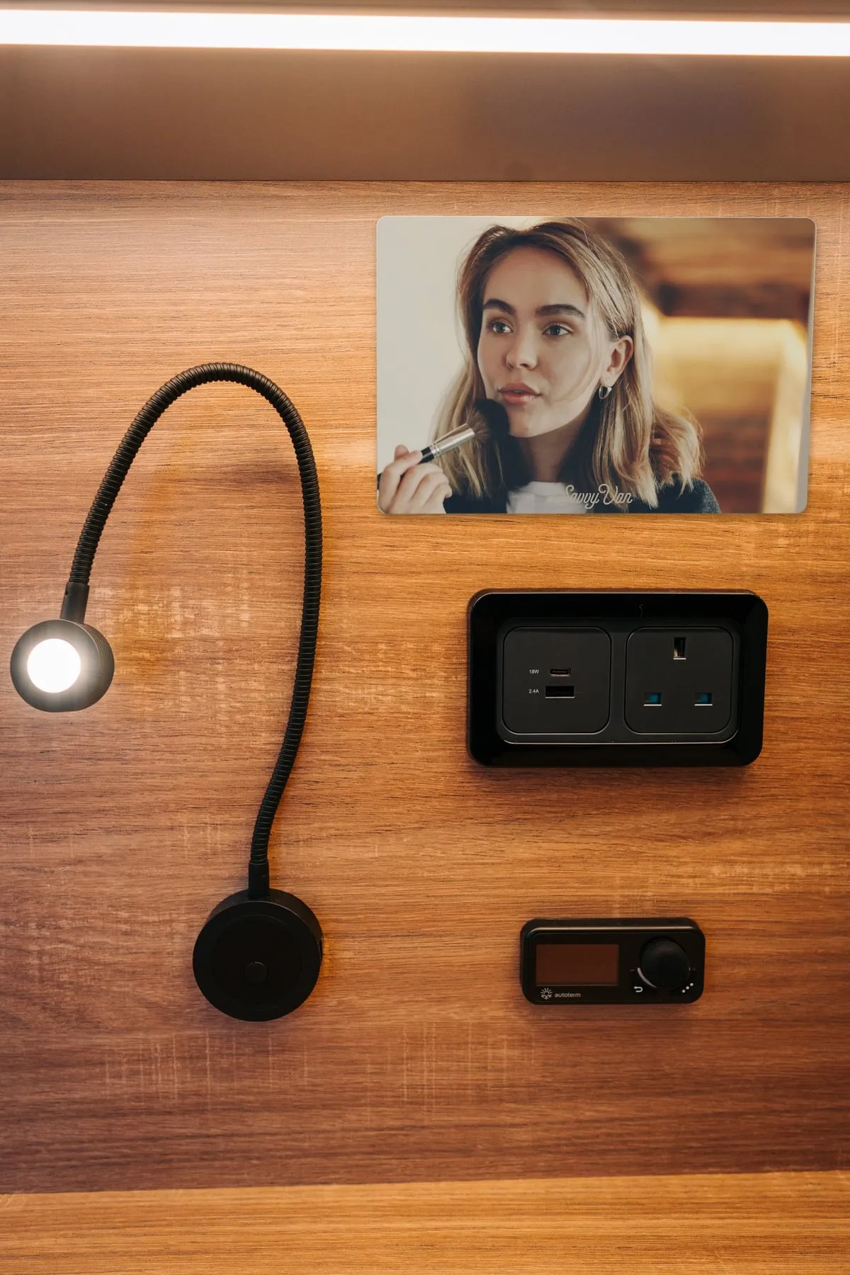 The mirrored screen switched off on a wood-panelled van wall, with a woman brushing on make-up reflected clearly in it, above a USB and mains socket and an Autoterm heater controller, beside a reading light