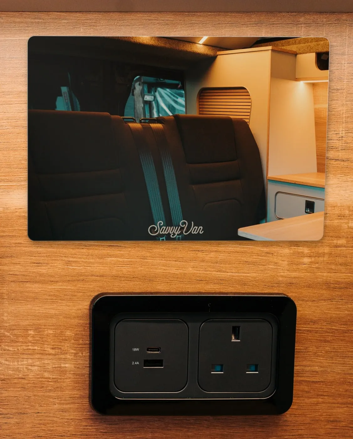 The mirrored screen switched off on a wood-panelled van wall above a USB and mains socket: a mirror reflecting the van's rear seats and kitchen, with the SavvyVan name small along its bottom edge