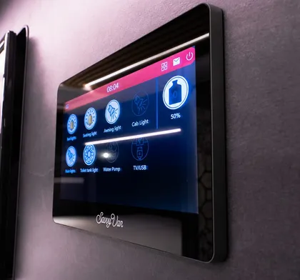 A 7 inch SavvyVan3 screen mounted on a grey soft-trim wall, seen at an angle, its lighting and water controls lit against the dark glass