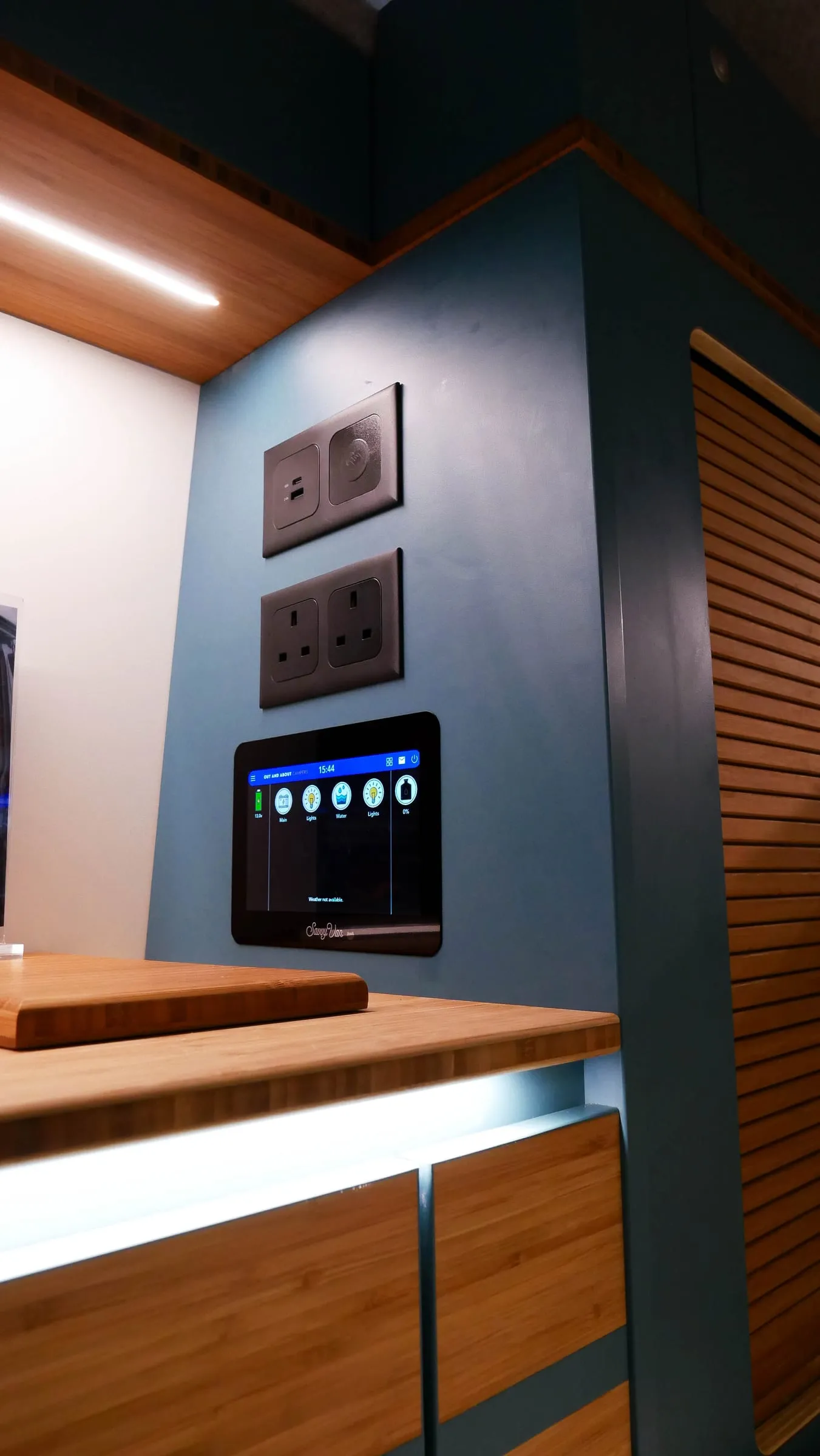 A SavvyVan3 panel fitted flush into a bamboo and teal campervan galley, screen showing lighting, water and power controls