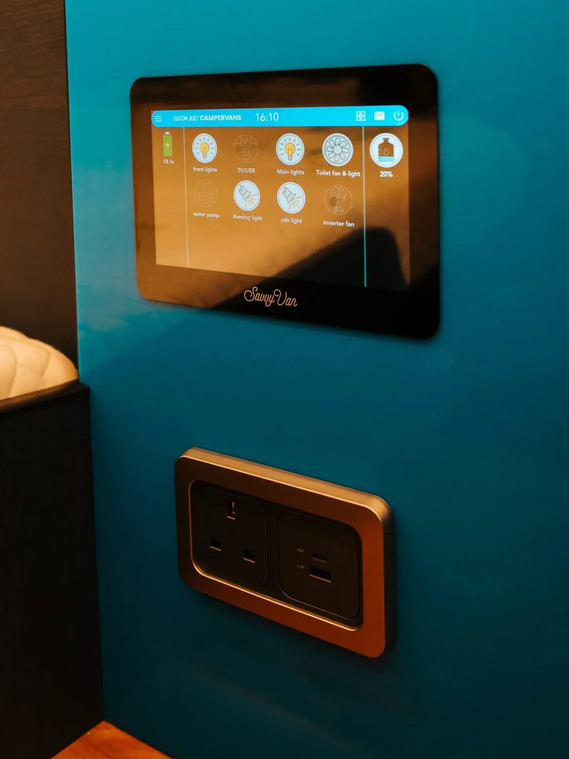 A SavvyVan3 panel fitted into a teal campervan wall above a double socket, its screen showing eight named lighting and power tiles and the fresh water level