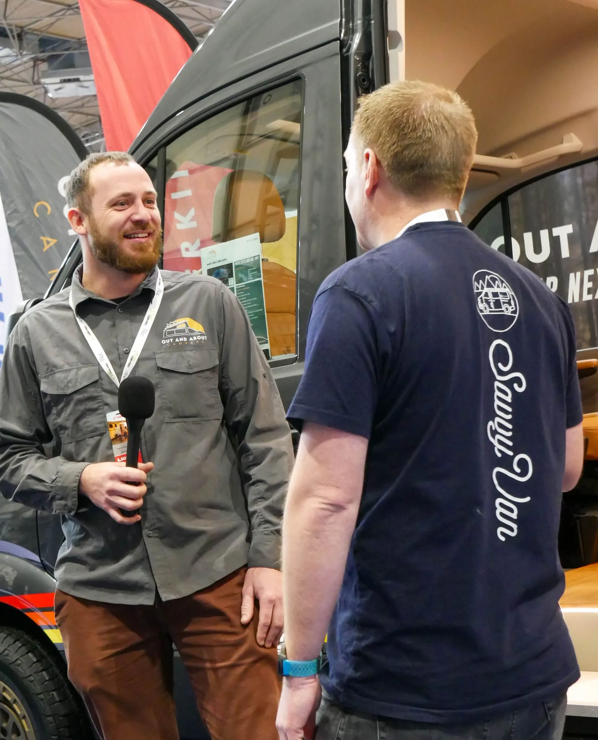 Simon from SavvyVan, in a SavvyVan t-shirt, talking with a member of the Out and About Campers team on their stand at a show, in front of one of their conversions