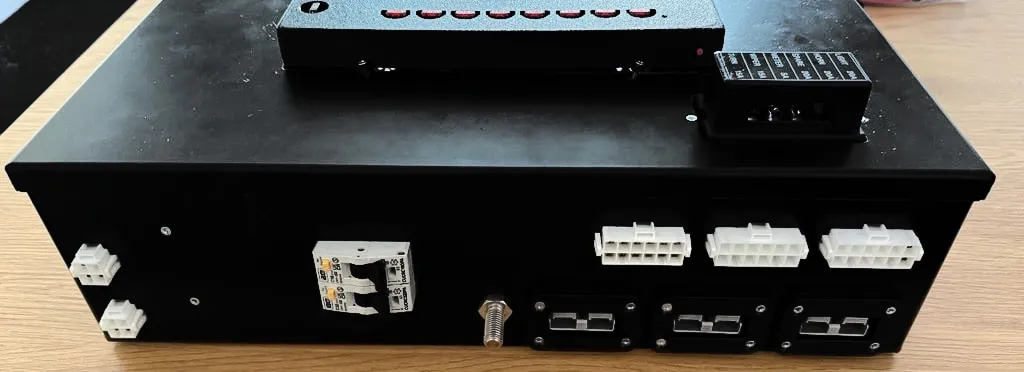 The connection side of SavvyCore: two small plugs, a pair of AC circuit breakers and a stud on the left, and three multi-pin loom plugs above three high-current connectors on the right, with the fuse panel on the lid behind.