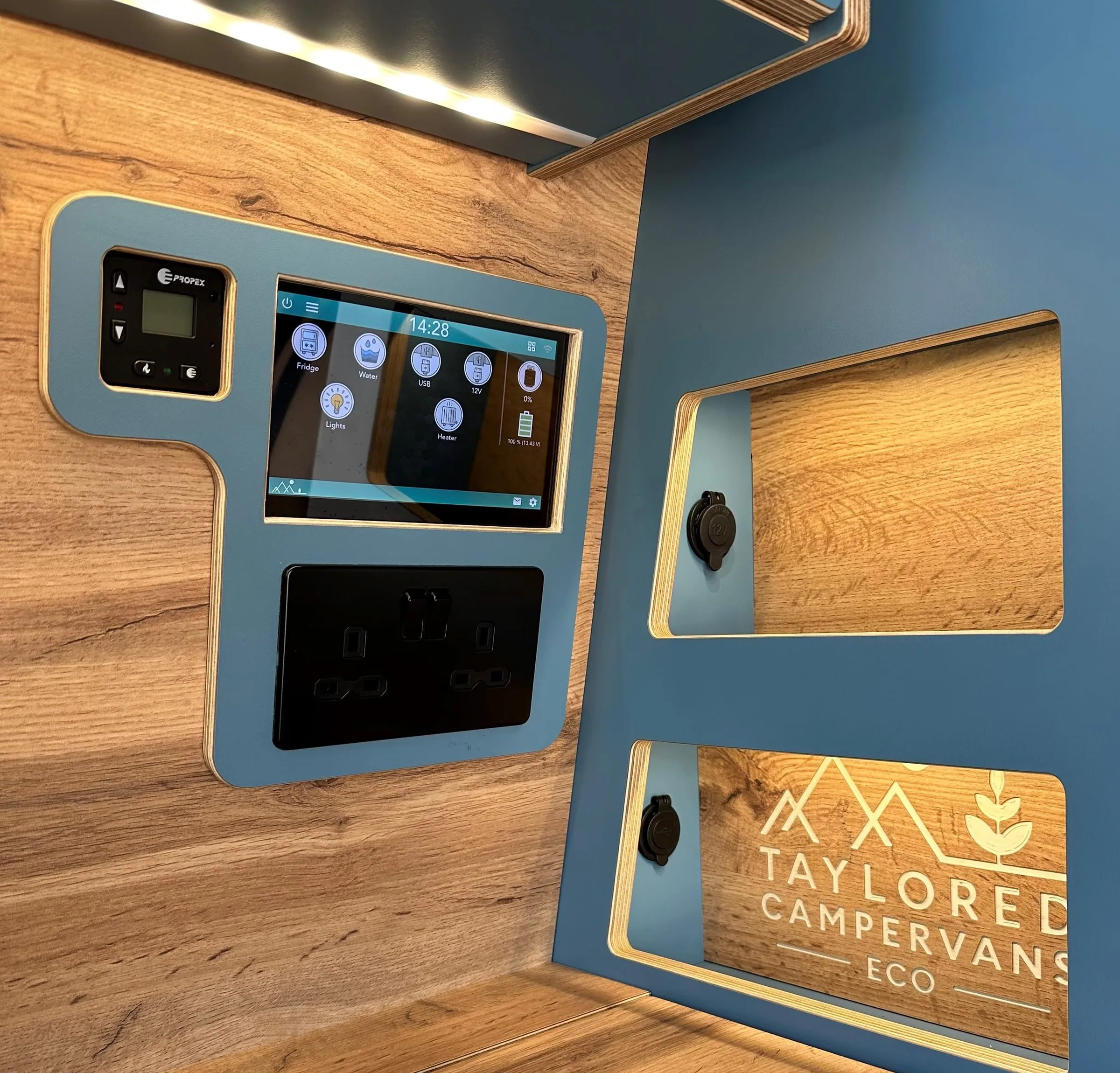 A SavvyVan Lite v2.1 panel fitted in a Taylored Campervans conversion, set into a blue surround beside a Propex heater control and above a double socket