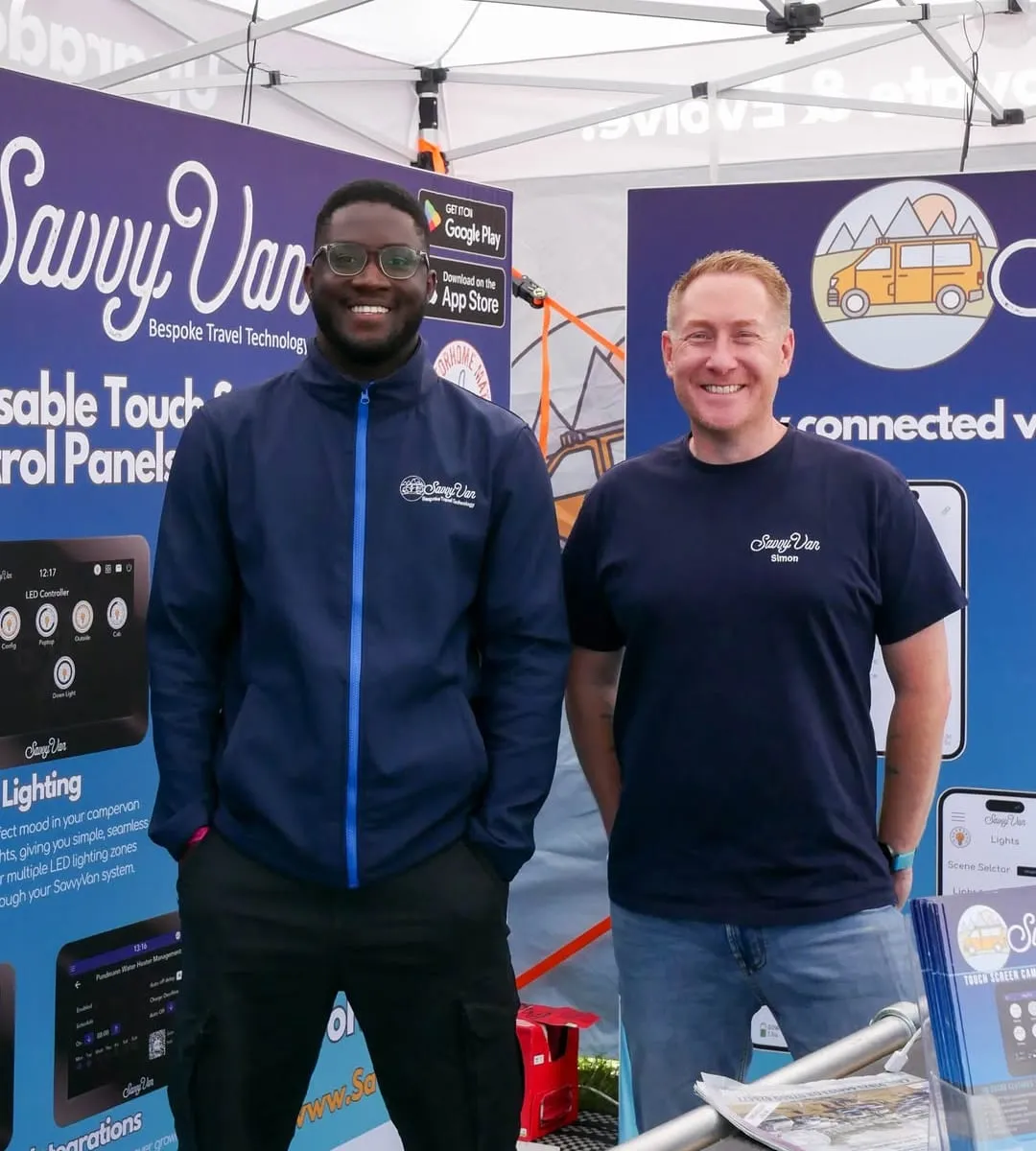 Parbi and Simon from SavvyVan smiling on the SavvyVan stand at the South West Motorhome and Campervan Show, with panel screens on the display boards behind them