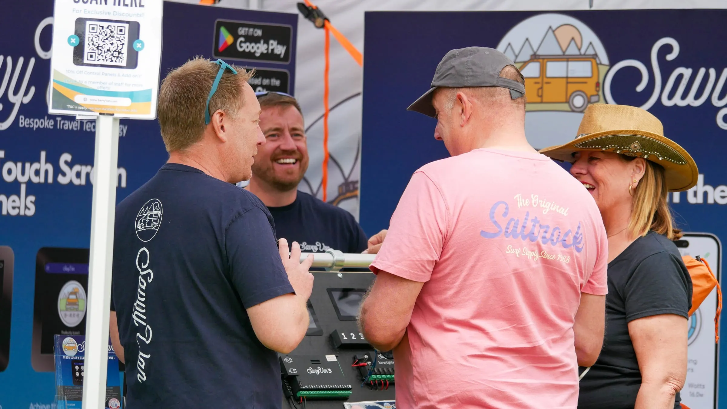 Two of the SavvyVan team in branded t-shirts talking and laughing with two visitors at a show stand, a SavvyVan3 control board and touchscreen laid out on the demonstration bench between them