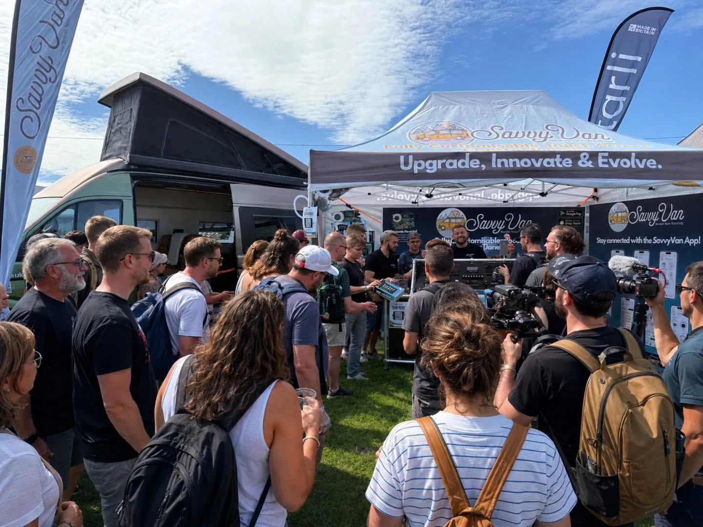 A crowd gathered around the SavvyVan stand at VanLife Festival in Shrewsbury, with a pop-top campervan open alongside it