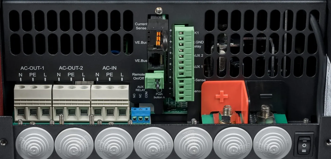 The connection area of a Victron MultiPlus-II with its front cover off: AC-OUT-1, AC-OUT-2 and AC-IN terminals on the left, a column of small connectors in the middle, the red-covered battery terminals on the right, and the on/off rocker switch at the bottom right