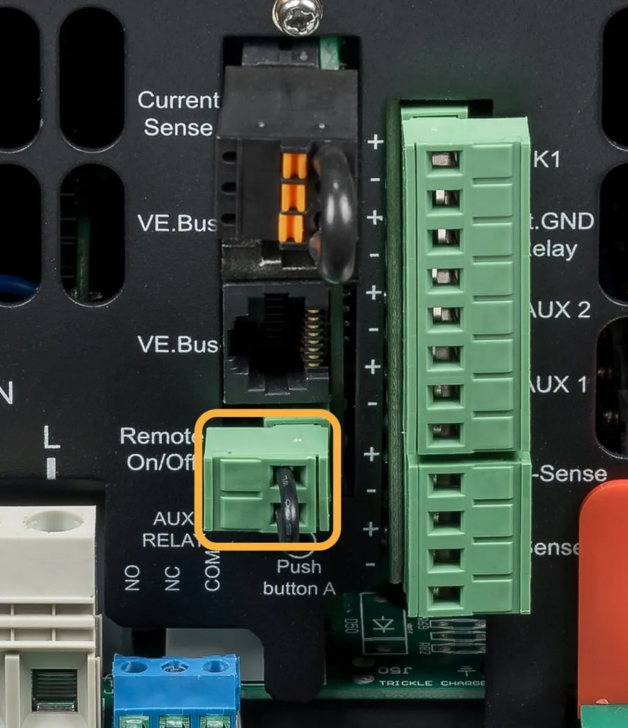 A close-up of the MultiPlus-II's middle connectors. The green two-way plug labelled Remote On/Off is ringed in orange, with a short black wire link looped across its two terminals. Below it, a blue three-way terminal is labelled AUX RELAY, NO, NC, COM